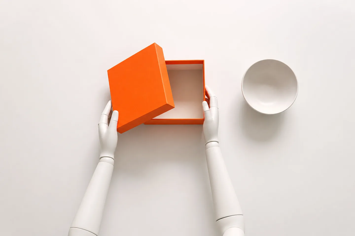 Two white mannequin hands seen from above lifting the lid off an orange box, next to an empty white bowl